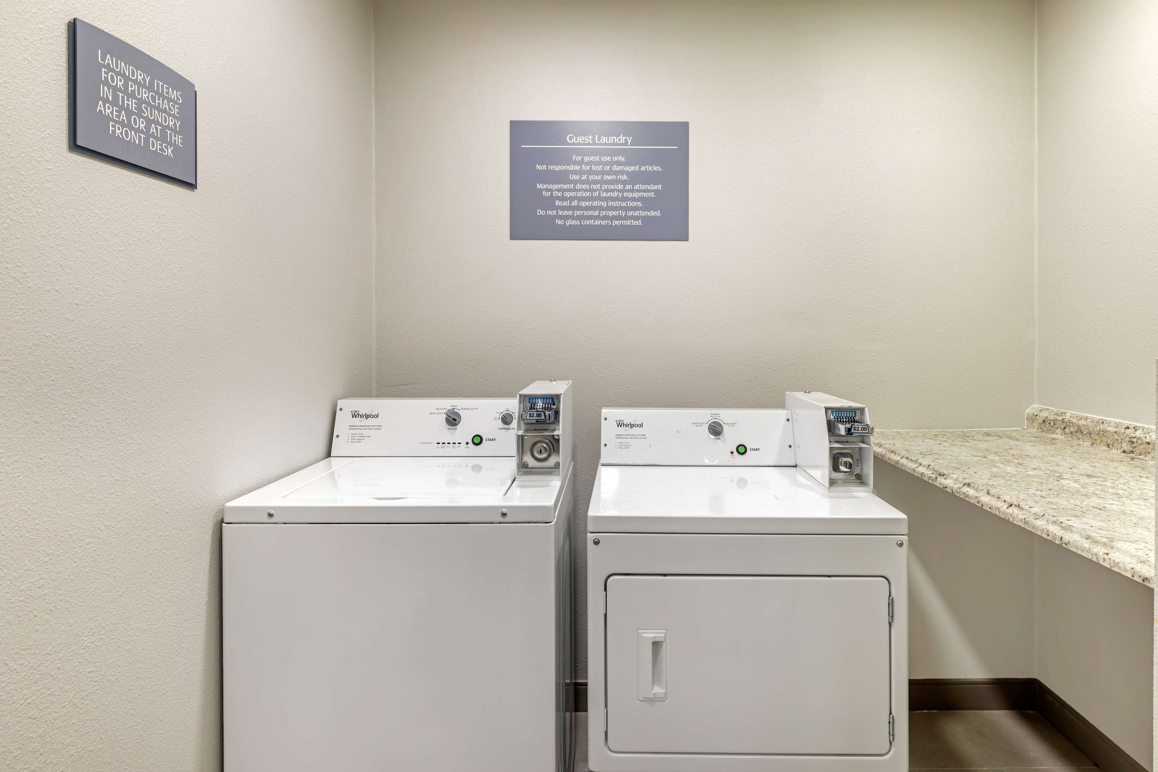 A laundry area with two Whirlpool washers and dryers, a sign indicating guest laundry rules, and a countertop.