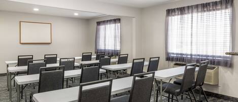 Un salón de conferencias con mesas blancas y sillas grises, ventanas amplias con cortinas y un cuadro en la pared.