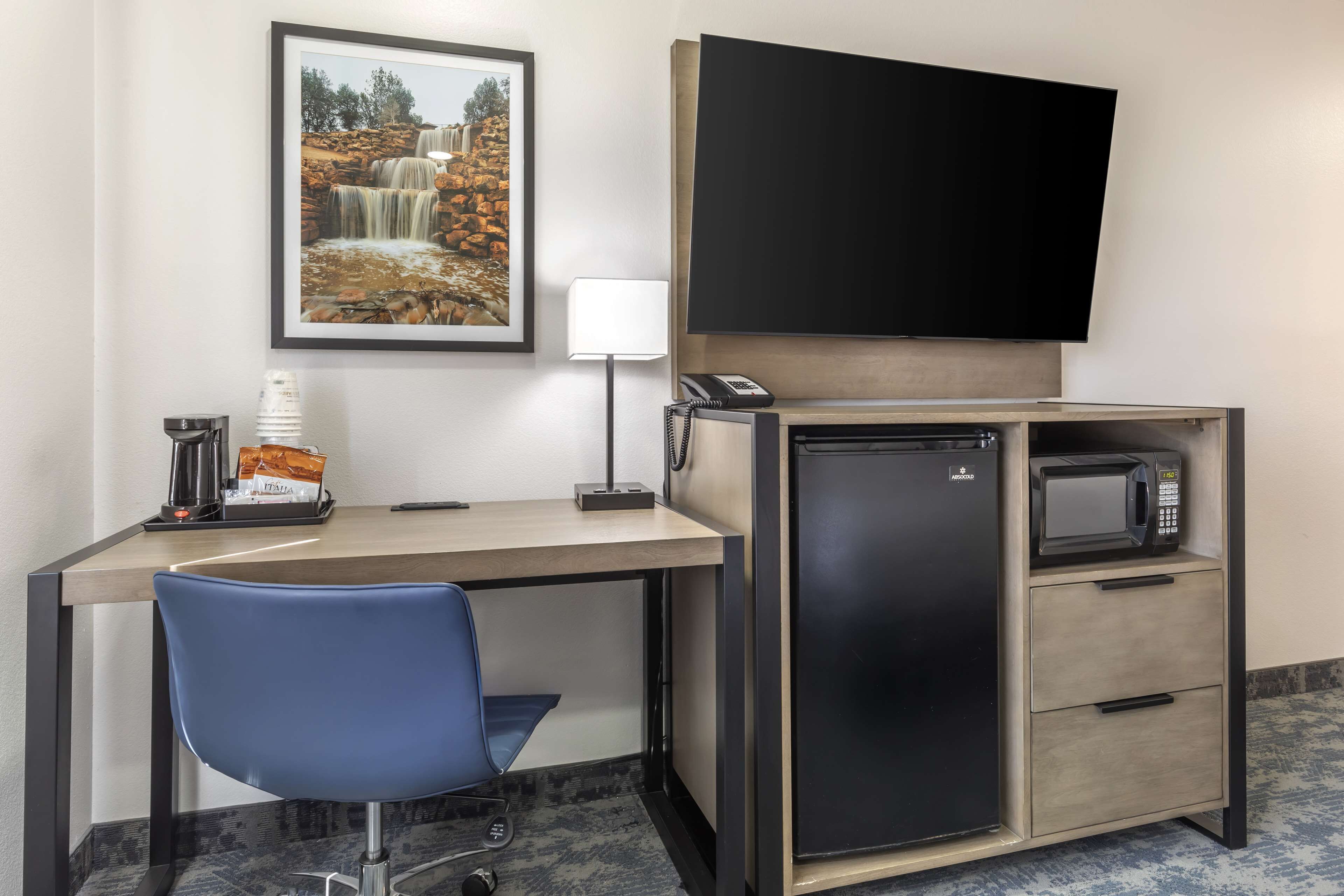 A hotel room with a desk, a chair, a refrigerator, a microwave, a telephone, a coffee maker, a lamp, and a framed picture of a waterfall.