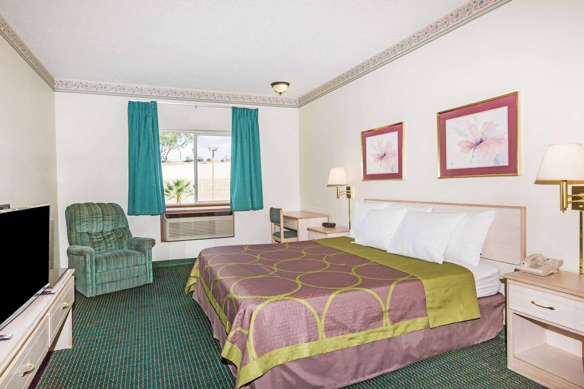 A hotel room with a bed, a chair, a desk, a lamp, a TV, and two framed pictures on the wall.