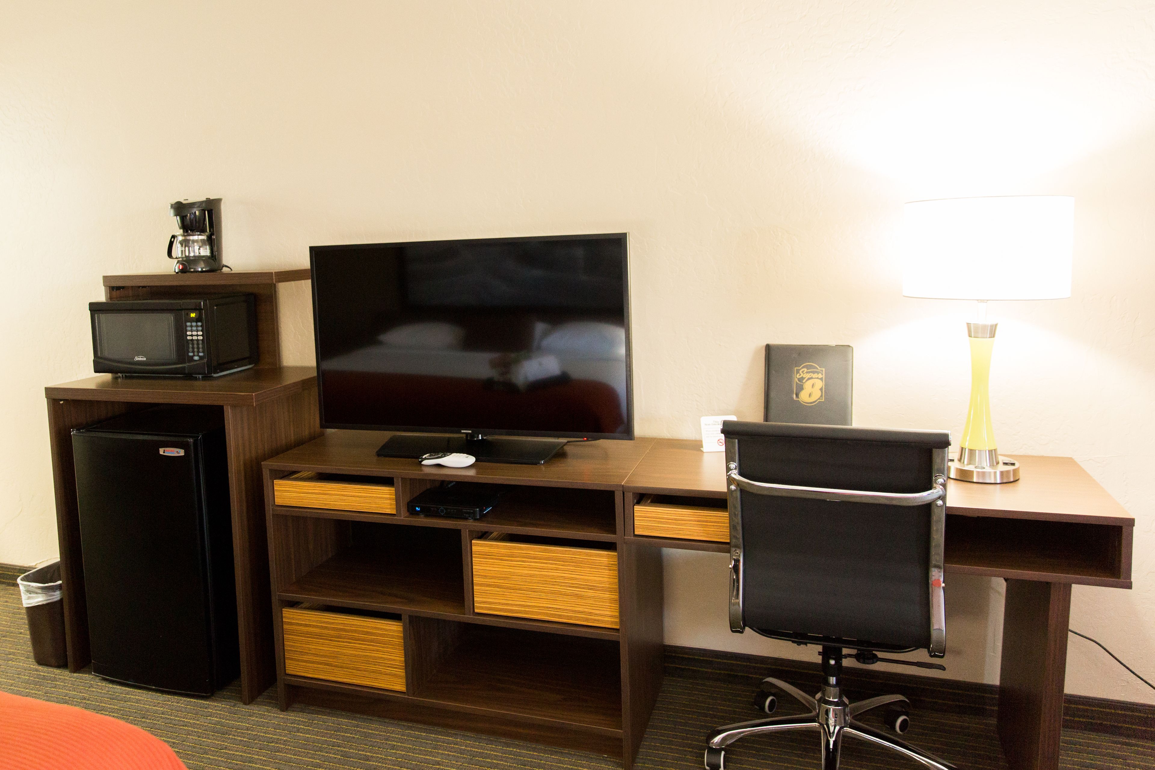 A hotel room with a wooden desk, a television, a microwave, a refrigerator, a chair, and a desk lamp.