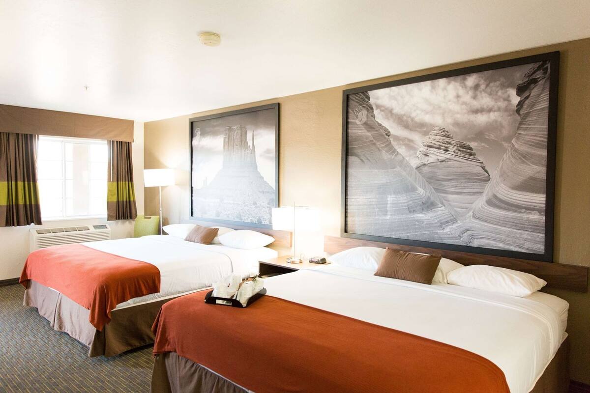 Une chambre d’hôtel avec deux lits, une grande fenêtre avec des rideaux, un climatiseur fixé au mur et deux grands tableaux encadrés.