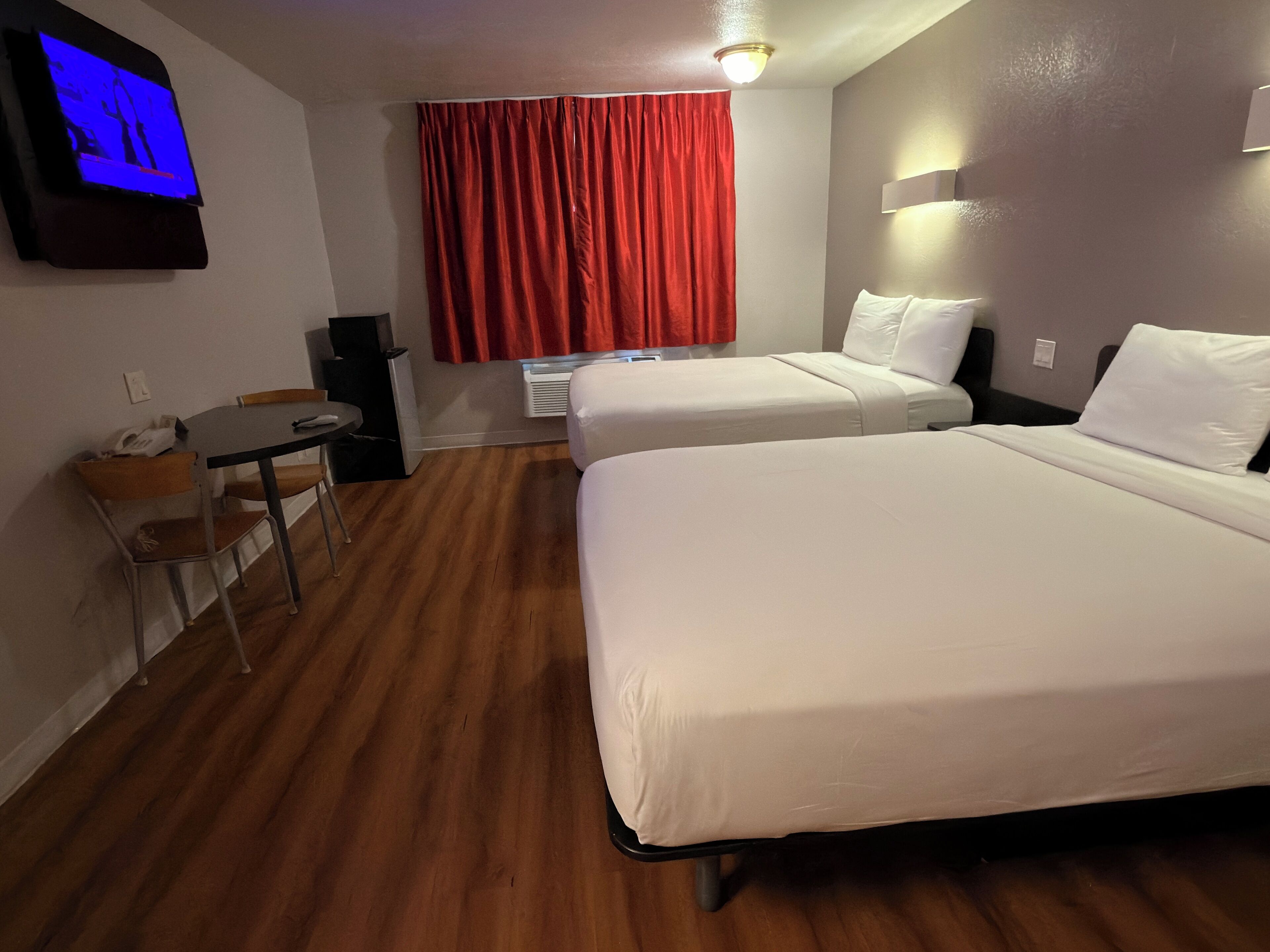 A hotel room with two beds, a desk with a chair, a TV mounted on the wall, and a red curtain.