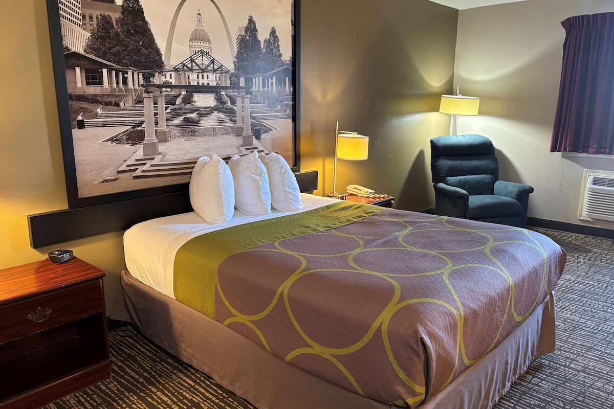 A hotel room with a large bed, a nightstand, a chair, a lamp, and a framed picture on the wall.