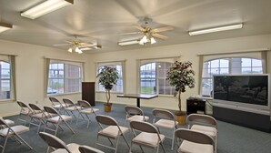 Un salón de conferencias con sillas plegables, una mesa, ventiladores de techo, ventanas amplias y una pantalla de televisión.