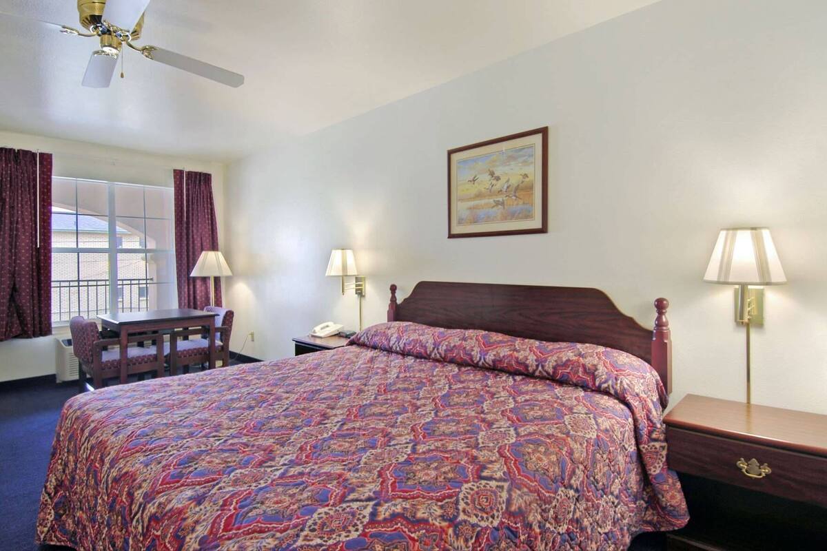 Une chambre d’hôtel comprenant un lit, un bureau, une chaise, deux lampes de chevet, un ventilateur de plafond et un tableau au mur.