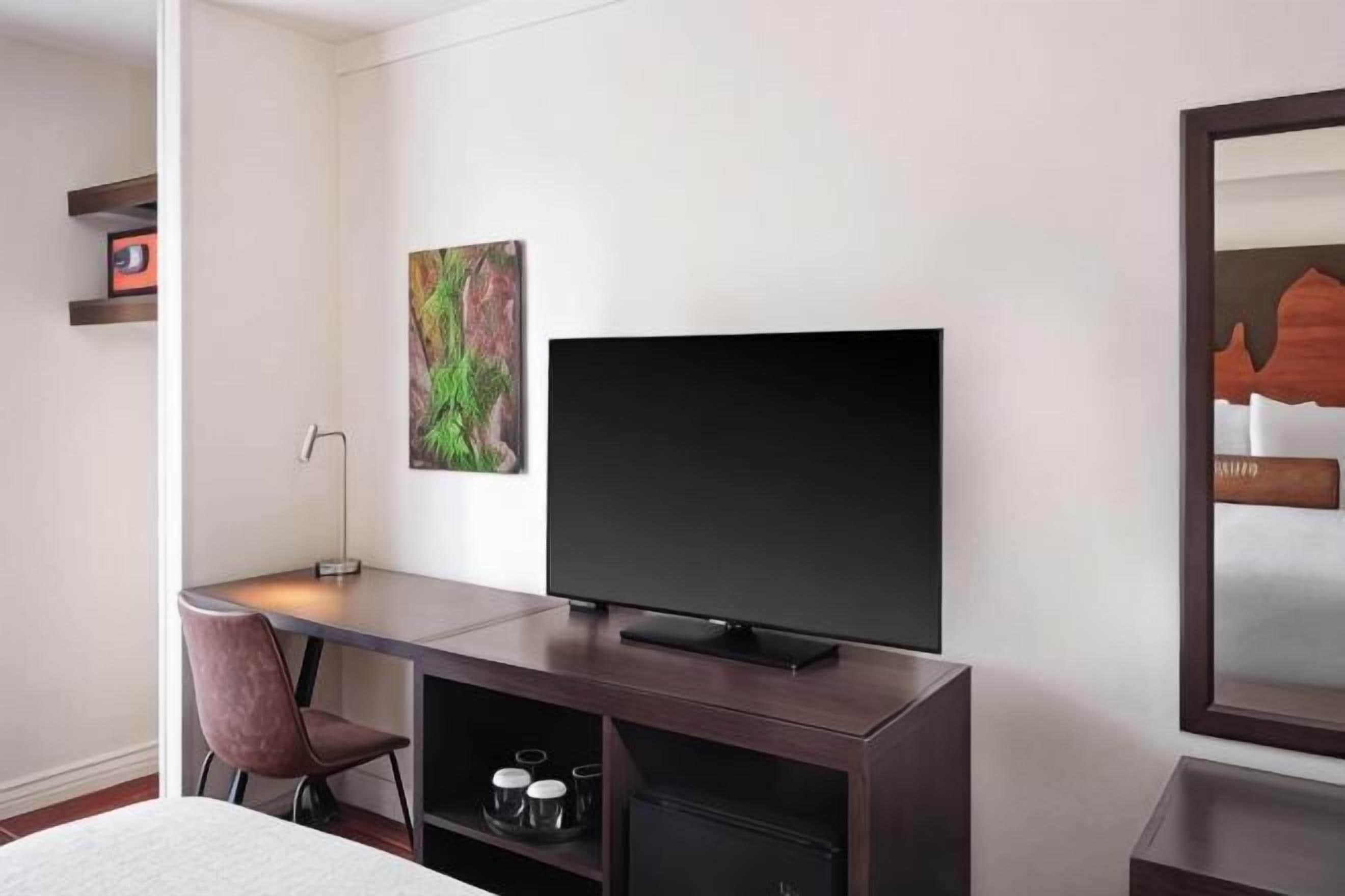 Ein Hotelzimmer mit Fernseher auf einem Holztisch, einem Sessel, einem Bett und einem Bild an der Wand.