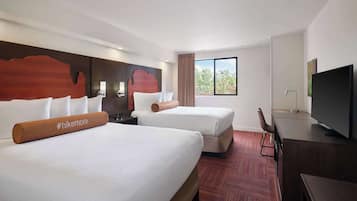 A hotel room with two beds, a TV, a desk, and a window with a view of trees.