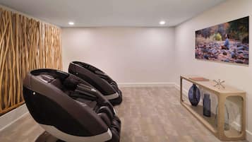 A massage therapy room with two massage chairs, a wooden wall panel, a shelf with decorative items, and a painting of a natural scene.