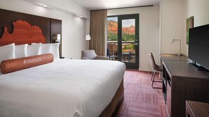 Habitación de hotel con una cama grande, un escritorio, un televisor y un balcón con vistas a formaciones rocosas rojas.