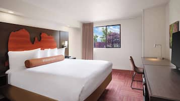 Ein Hotelzimmer mit einem großen Bett, einem Schreibtisch, einem Sessel, einem Fernseher und einem Fenster mit Blick auf Bäume.