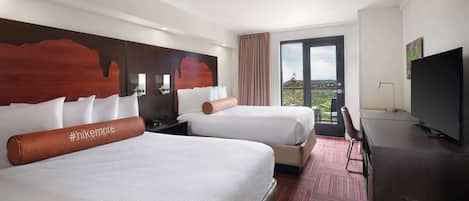 Habitación de hotel con dos camas, un escritorio, televisión y balcón con vistas.