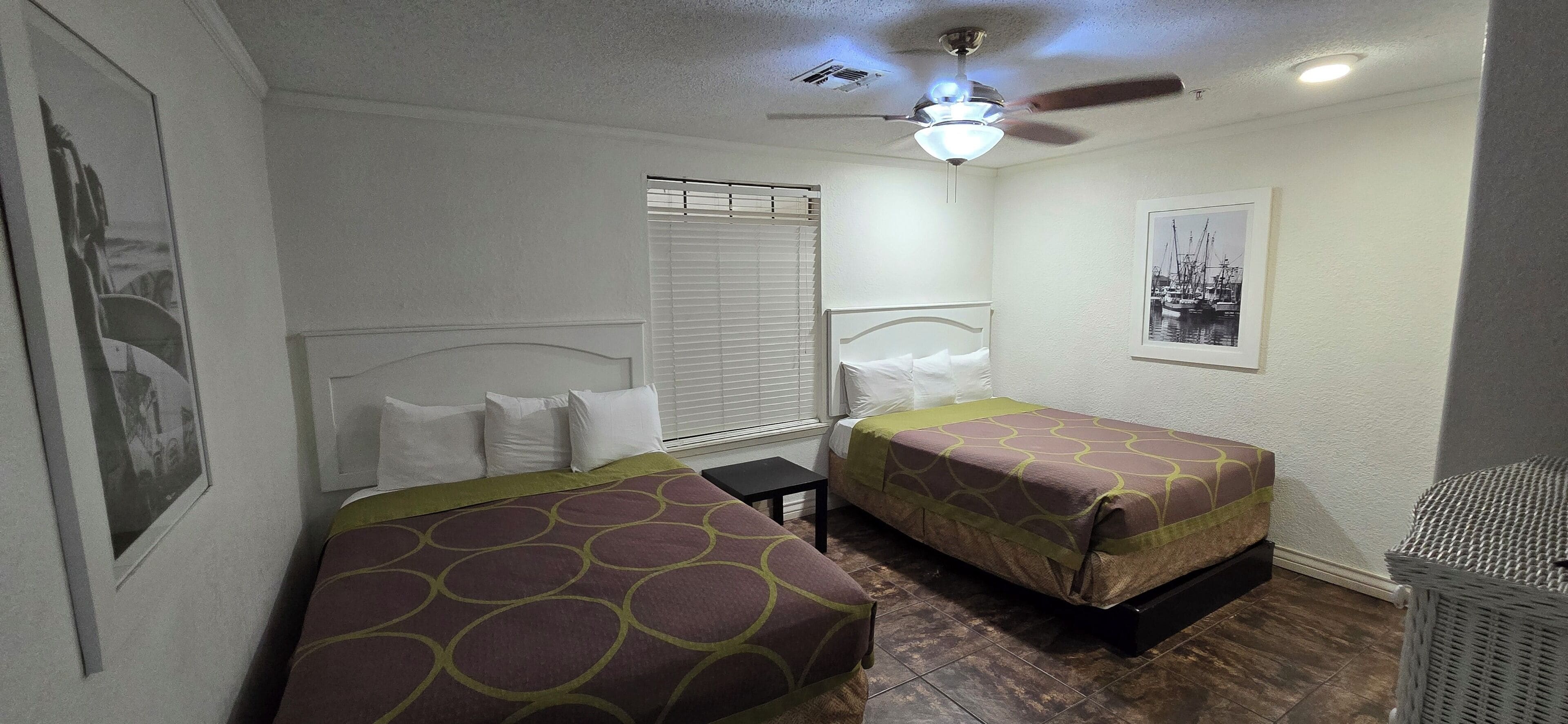 A hotel room with two single beds, a ceiling fan, a window with blinds, and a framed picture on the wall.
