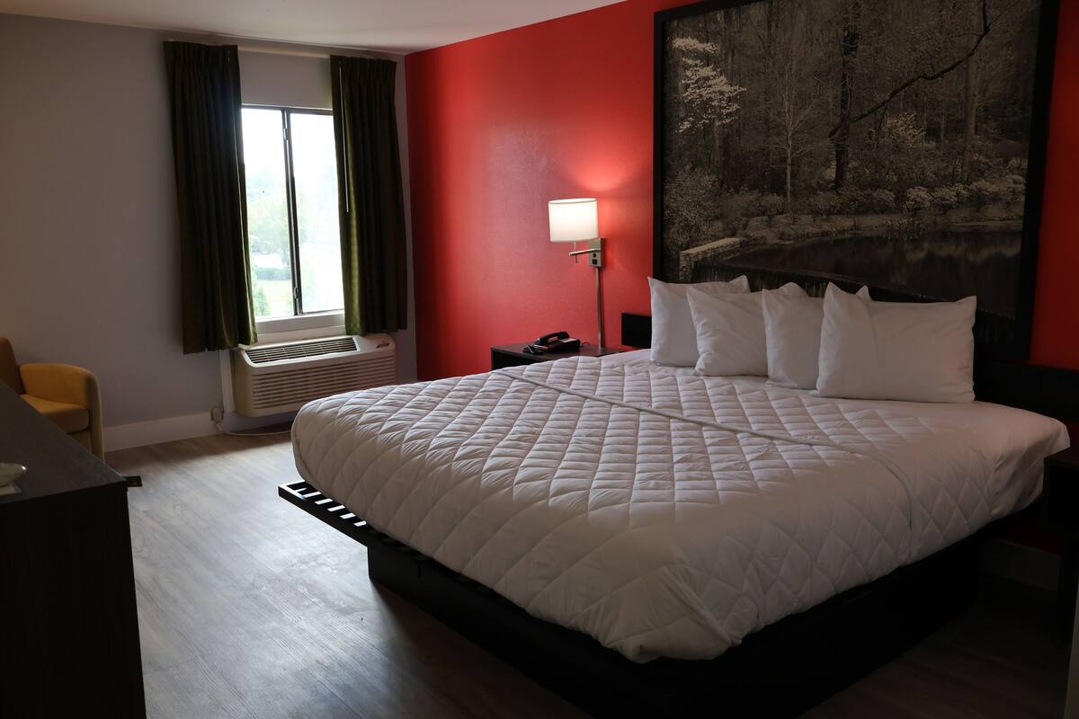 Habitación de hotel con una cama grande, un escritorio, una silla, un ventanal con cortinas, una lámpara de pared y un cuadro.
