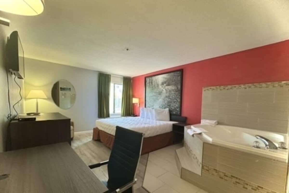 Habitación de hotel con cama, escritorio, silla, lámpara de escritorio, televisor montado en la pared, baño con bañera y un amplio ventanal con cortinas.