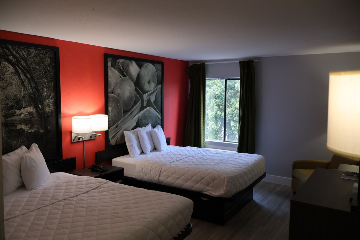 Ein Hotelzimmer mit zwei Betten, einem großen abstrakten Gemälde an der Wand, einem Fenster mit Vorhängen und einem Schreibtisch mit einer Lampe.
