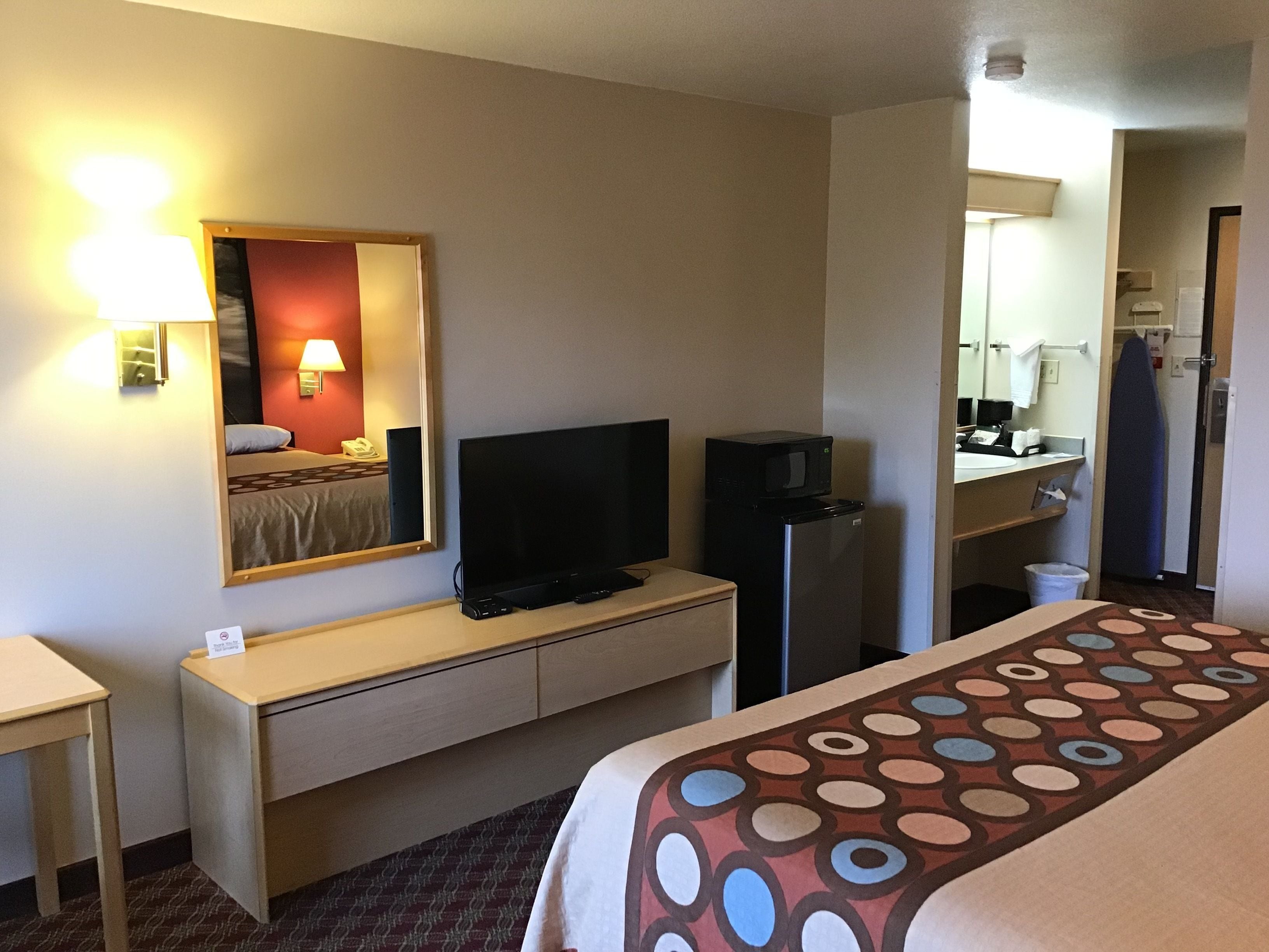 Une chambre d’hôtel équipée d’un lit, d’un bureau, d’une télévision, d’un réfrigérateur et d’une salle de bain, le tout visible à travers une porte ouverte.