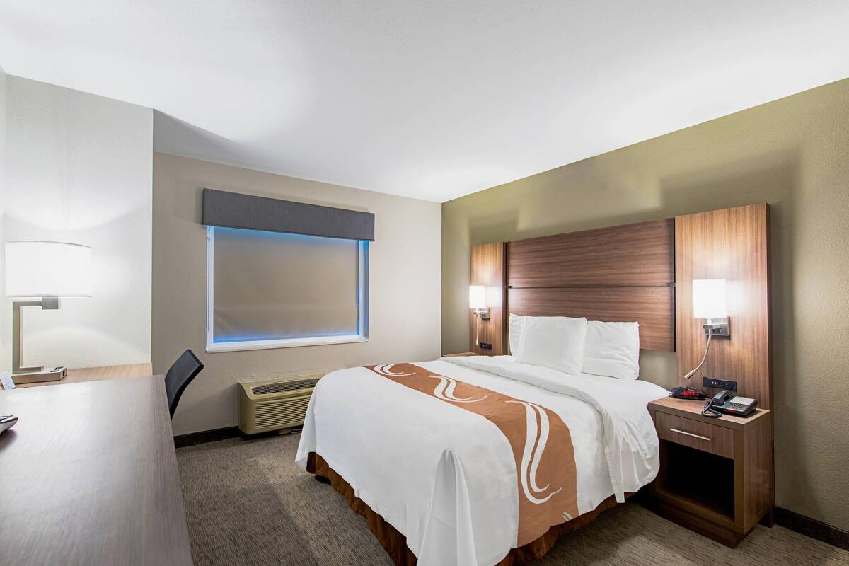 A hotel room with a bed, bedside tables, a desk, a lamp, a window with blinds, and a wall-mounted heater.