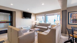 Una habitación de hotel moderna con un conjunto de sofá, un televisor montado en la pared y ventanas amplias que ofrecen vistas al exterior.