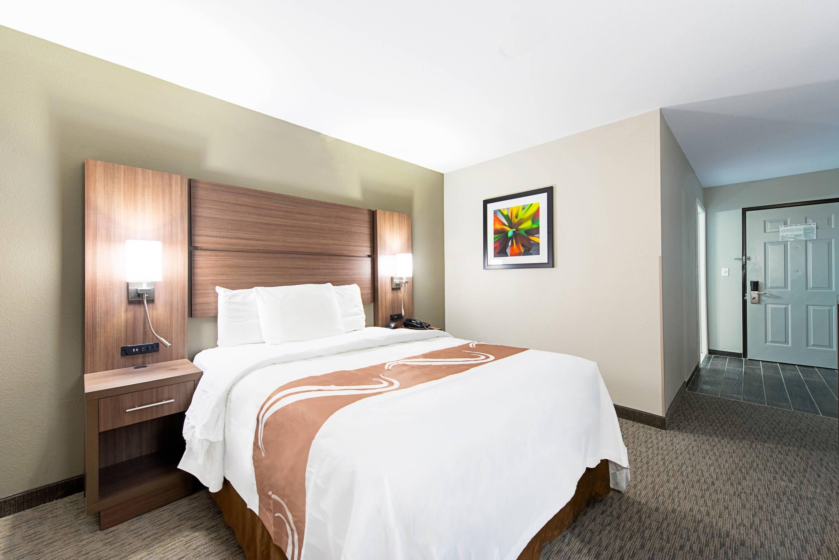 A hotel room with a bed, bedside tables, a nightstand, a wall-mounted lamp, a framed picture, and a door.