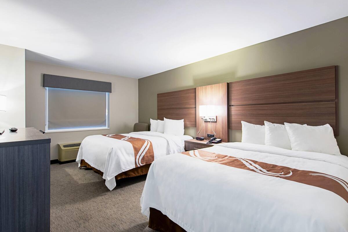 A hotel room with two beds, a desk, a window, and a wall-mounted lamp.