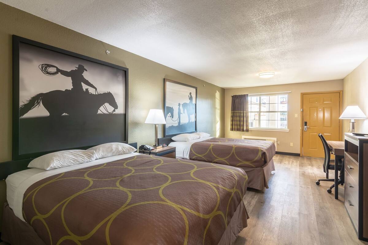 Une chambre d’hôtel avec deux lits, un bureau, une chaise, une fenêtre avec des rideaux et des œuvres d’art accrochées aux murs.
