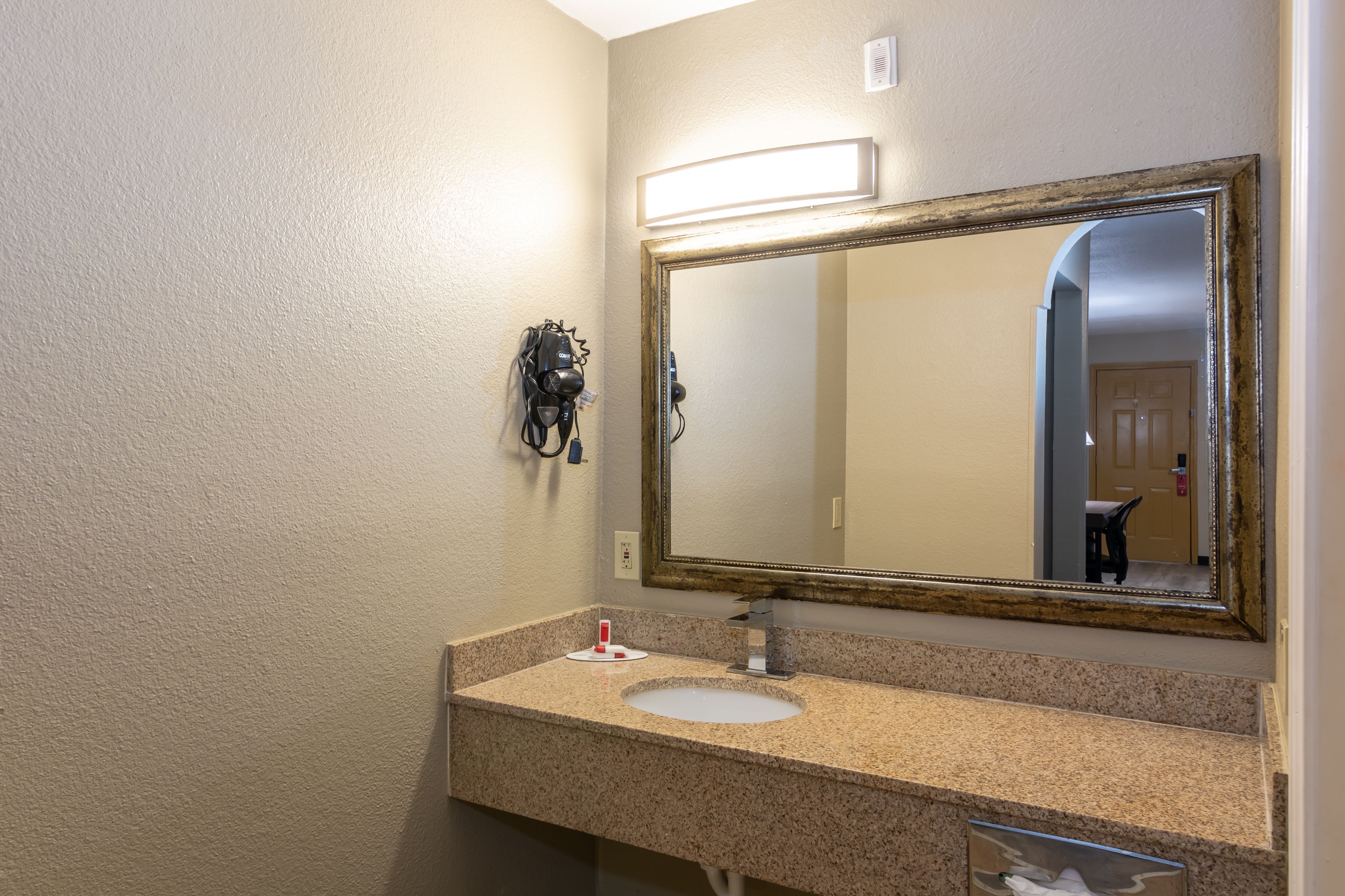 A bathroom with a granite countertop, a white sink, a large mirror, and a hairdryer on the wall.
