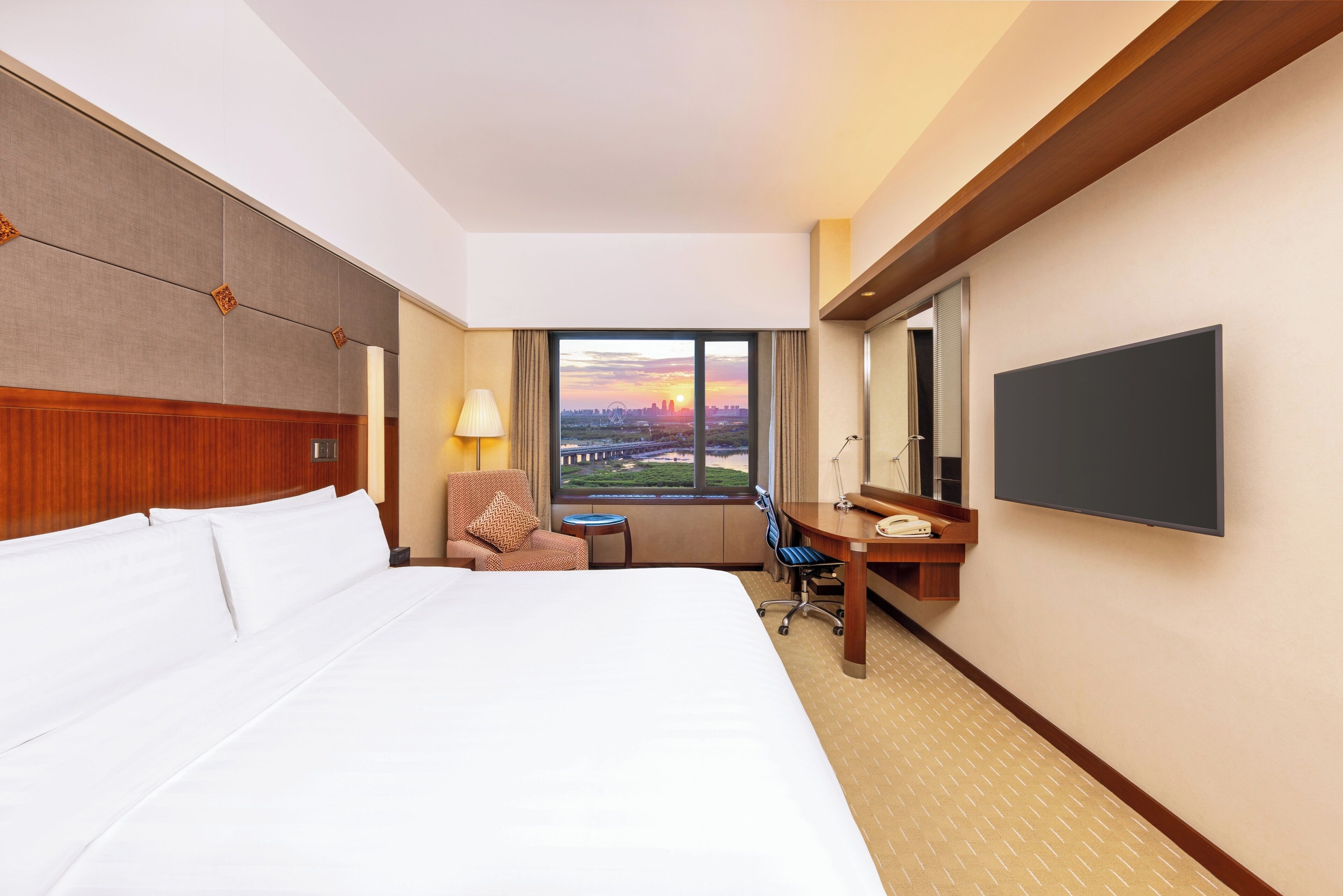 A hotel room with a large bed, a desk, a chair, a TV, and a window with a city view.