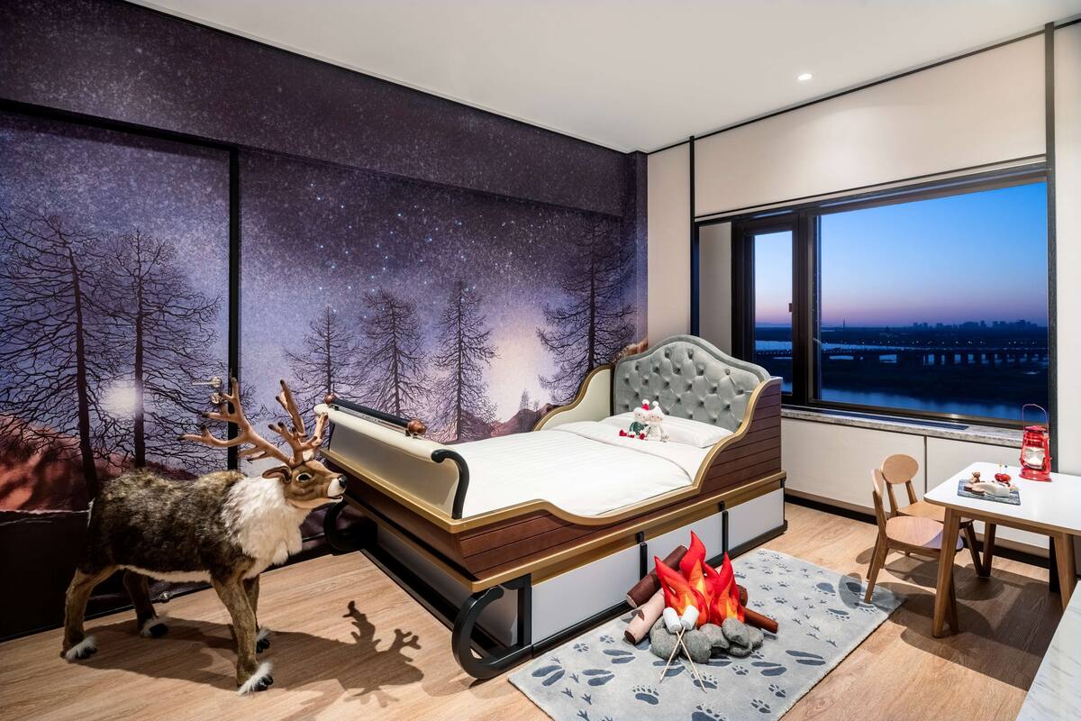 Un dormitorio con una cama, un mural del cielo nocturno, una estatua de un ciervo y vistas de un paisaje urbano a través de una ventana.