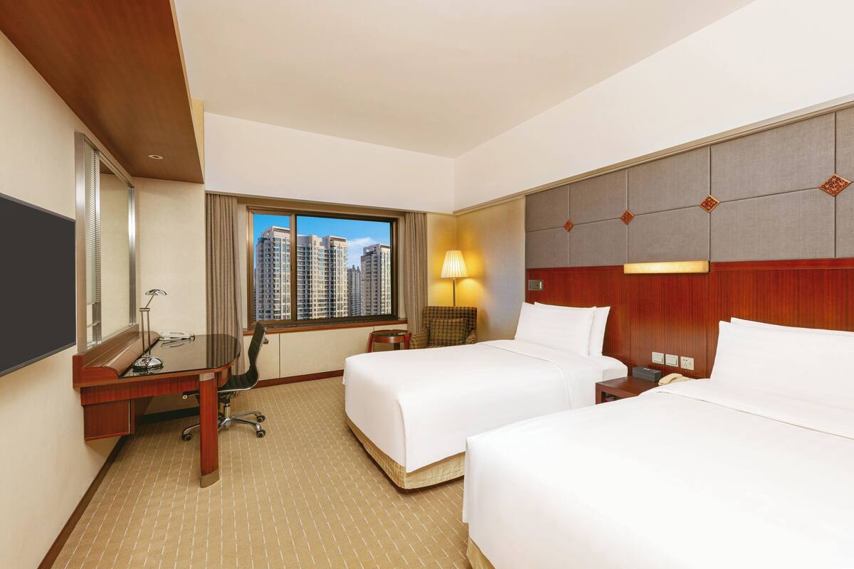Habitación de hotel con dos camas, un escritorio, una silla, una lámpara y vistas a edificios altos.