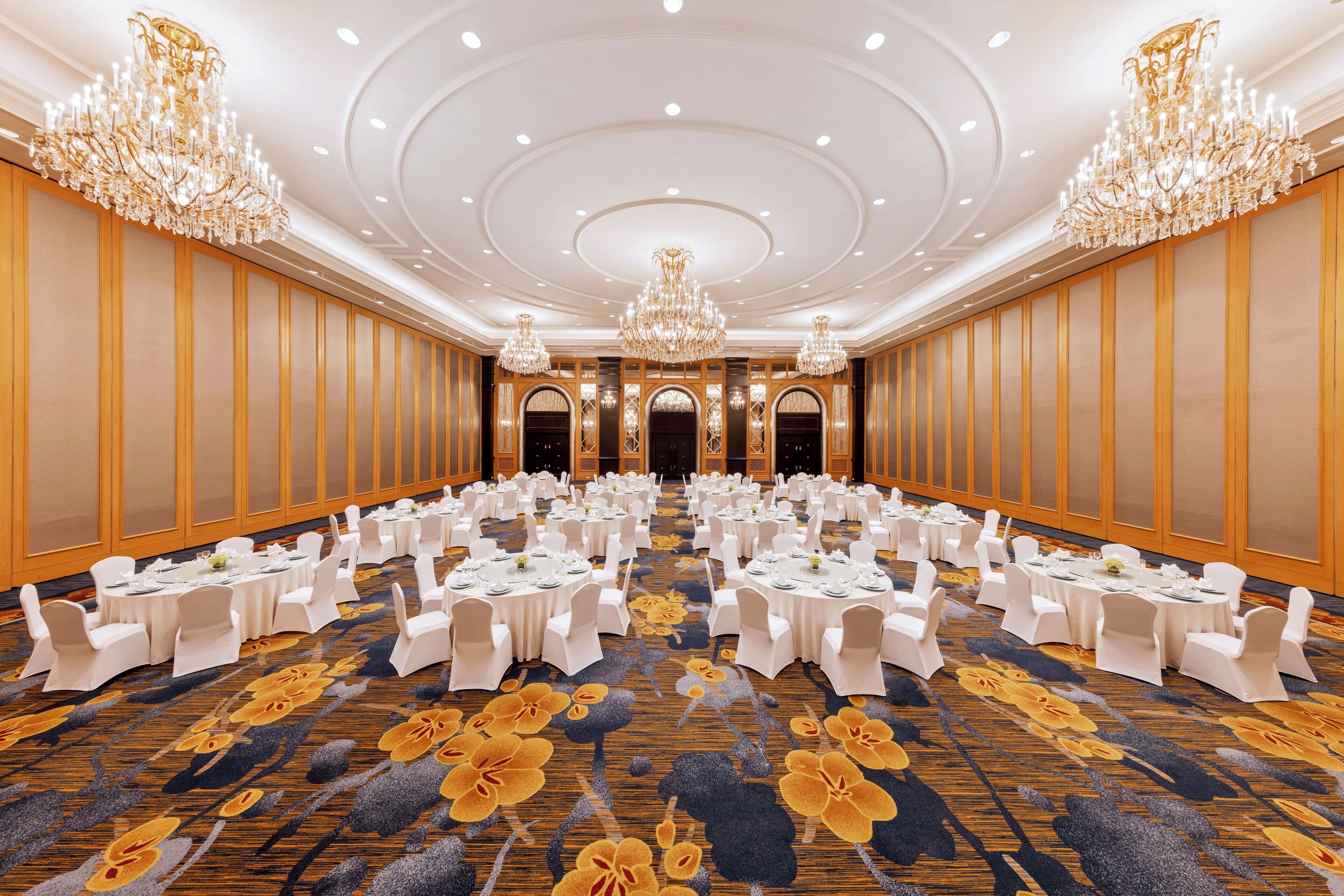 Un amplio salón de banquetes con mesas redondas, sillas blancas, candelabros y una alfombra floral.
