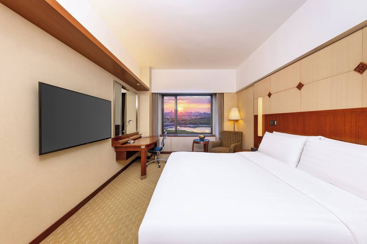 Habitación de hotel con una cama grande, un escritorio con silla, un televisor y una ventana con vista al atardecer.