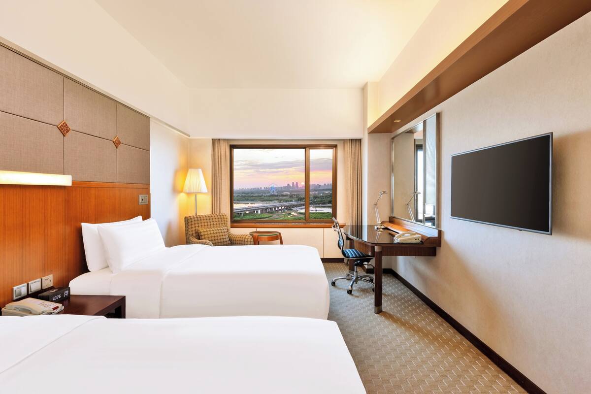 Habitación de hotel con dos camas, un escritorio, una silla, televisor y un ventanal con vista a la ciudad.