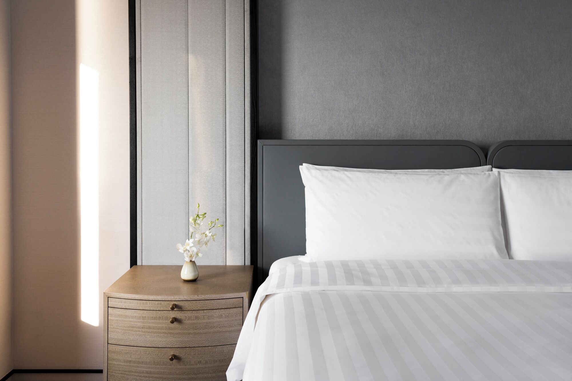 Een netjes opgemaakt bed met witte lakens en een grijs hoofdeinde, een houten nachtkastje met lades, en een vaas met witte bloemen.
