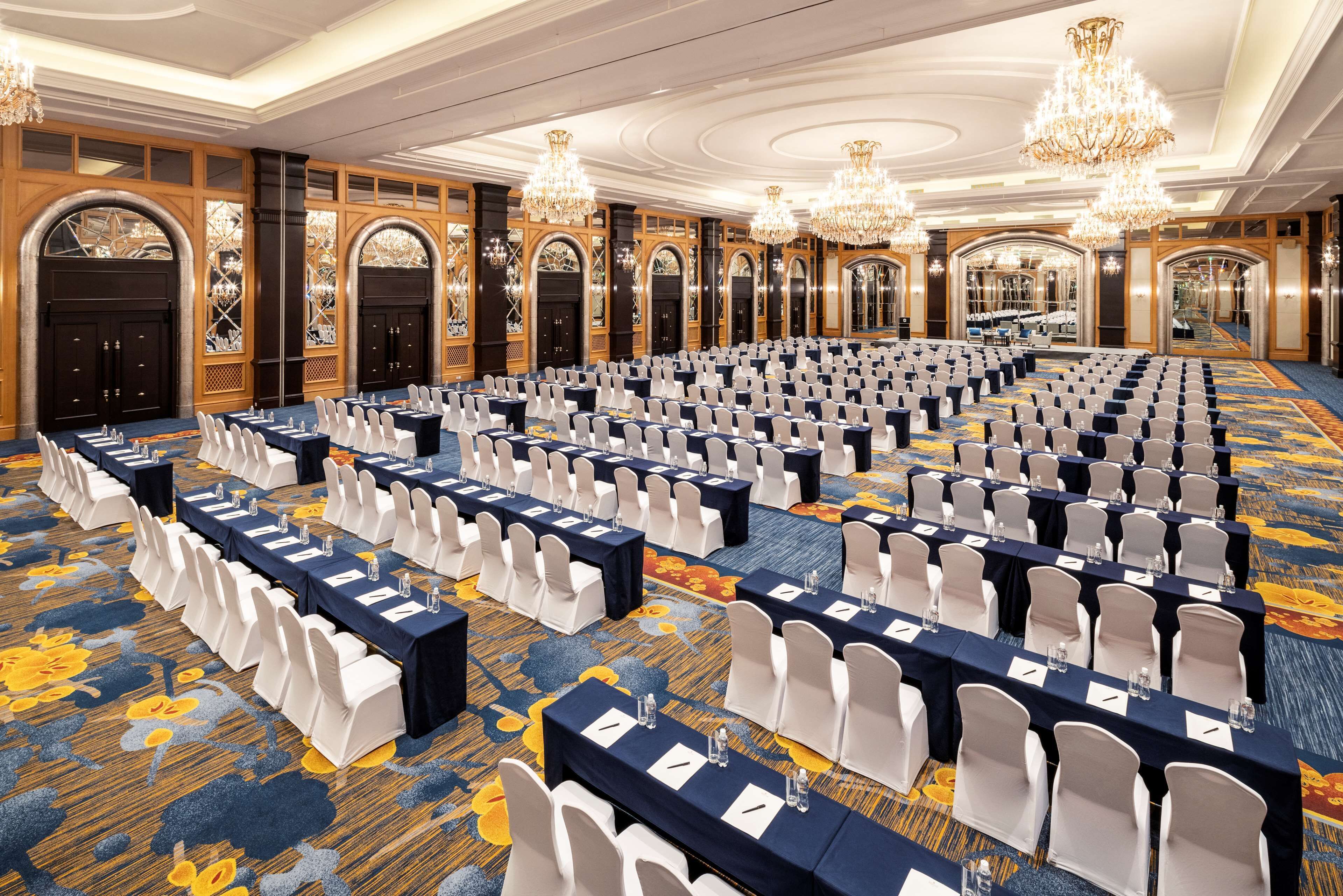 A large conference room with rows of tables and chairs, chandeliers, and ornate wooden doors.