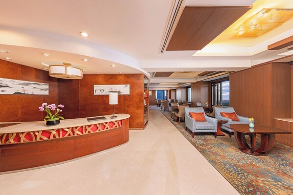 Zona de recepción de hotel con un mostrador de madera curvada, un área de descanso con sofás y sillones, y una alfombra estampada.