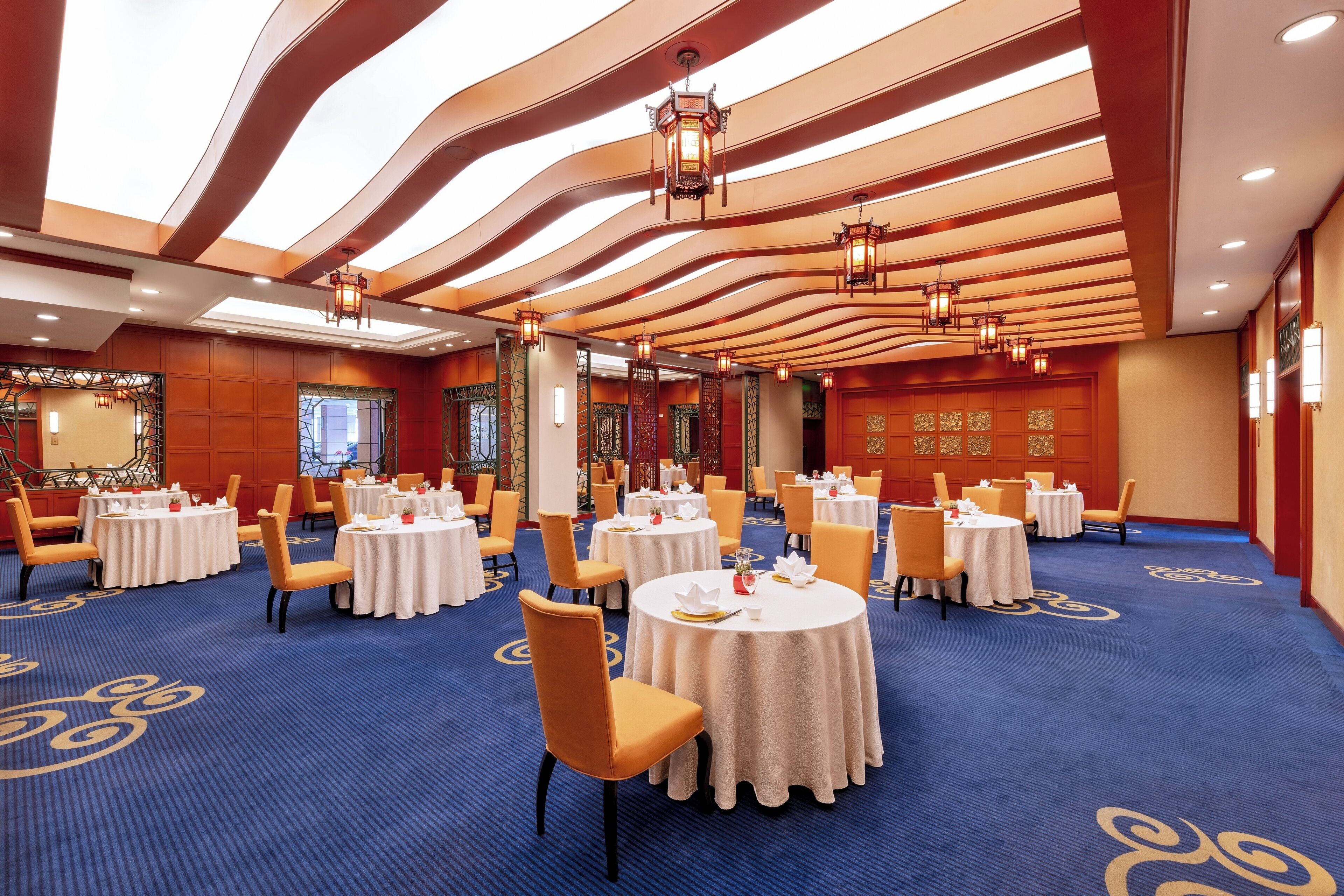 Une salle de banquet avec des tables rondes, des chaises et un tapis bleu orné d’un motif doré.