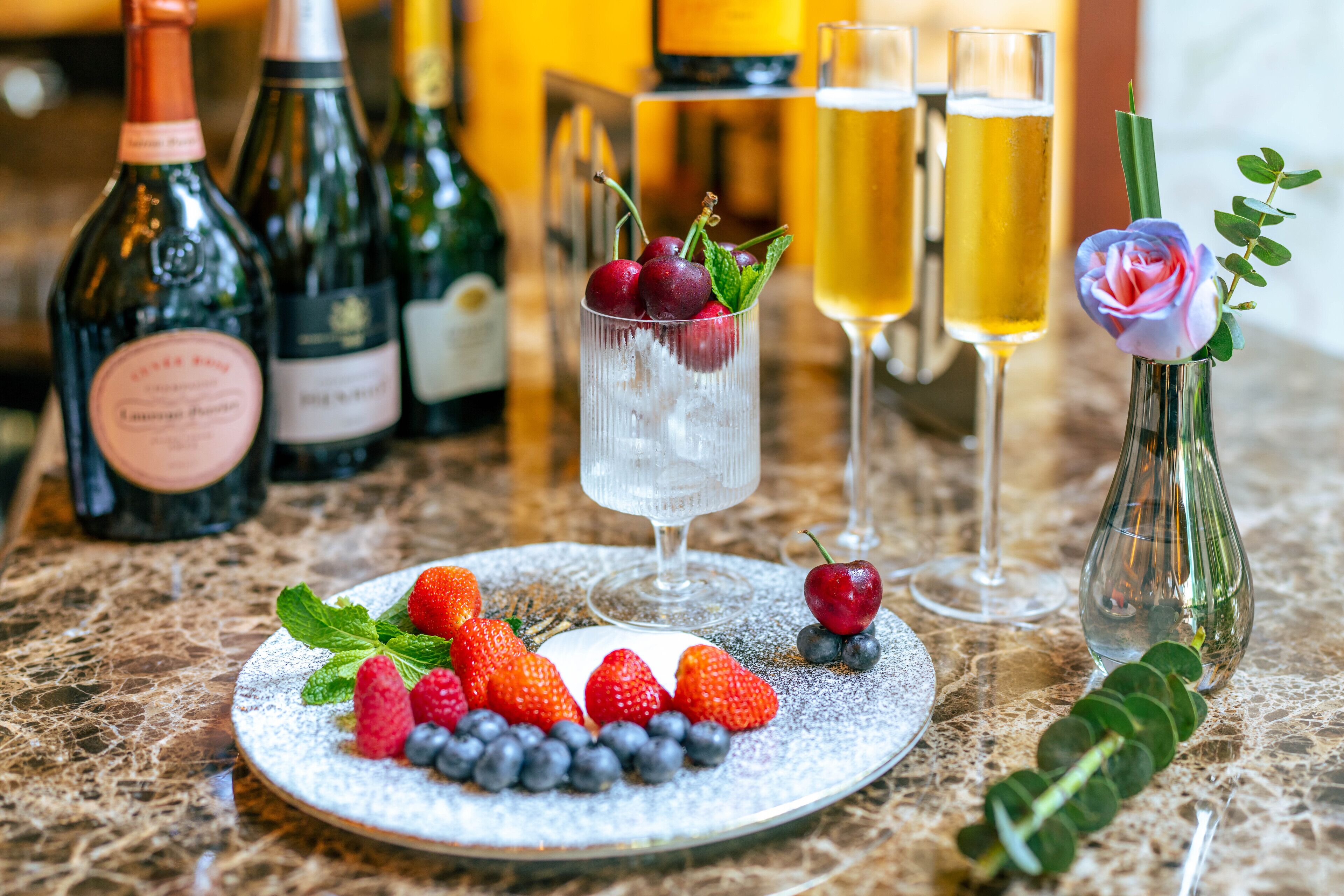 A table with a plate of strawberries, blueberries, and cherries, a glass of champagne, and bottles of wine.