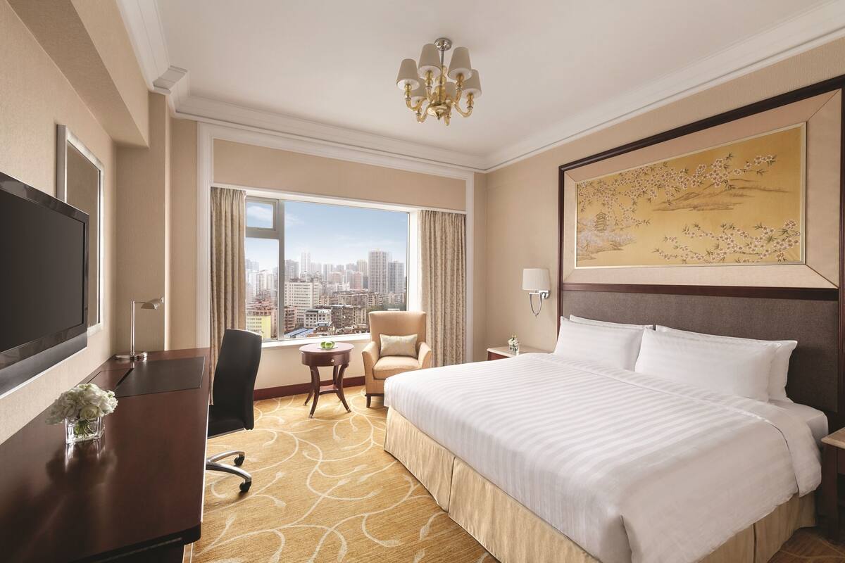 A hotel room with a large bed, a desk with a TV, a chair, a small table with a vase of flowers, and a large window with a city view.