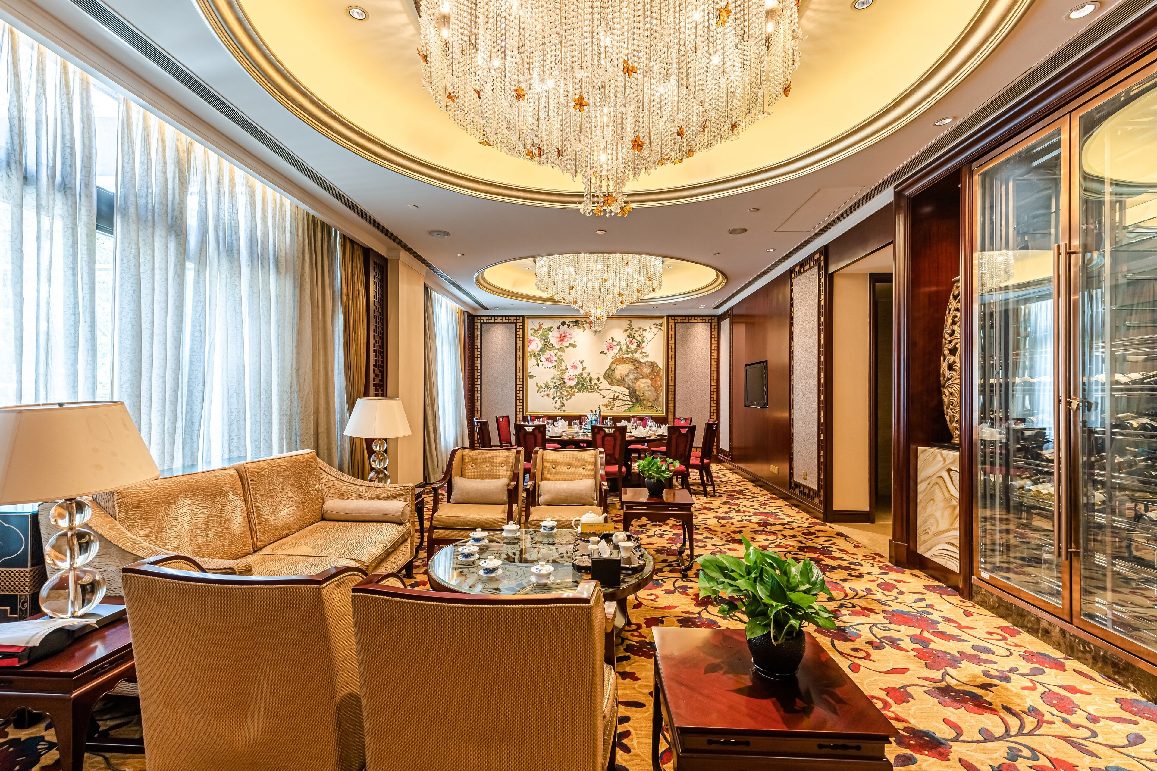A luxurious hotel room with a large chandelier, comfortable seating, a dining area, and a view of a cityscape through large windows.
