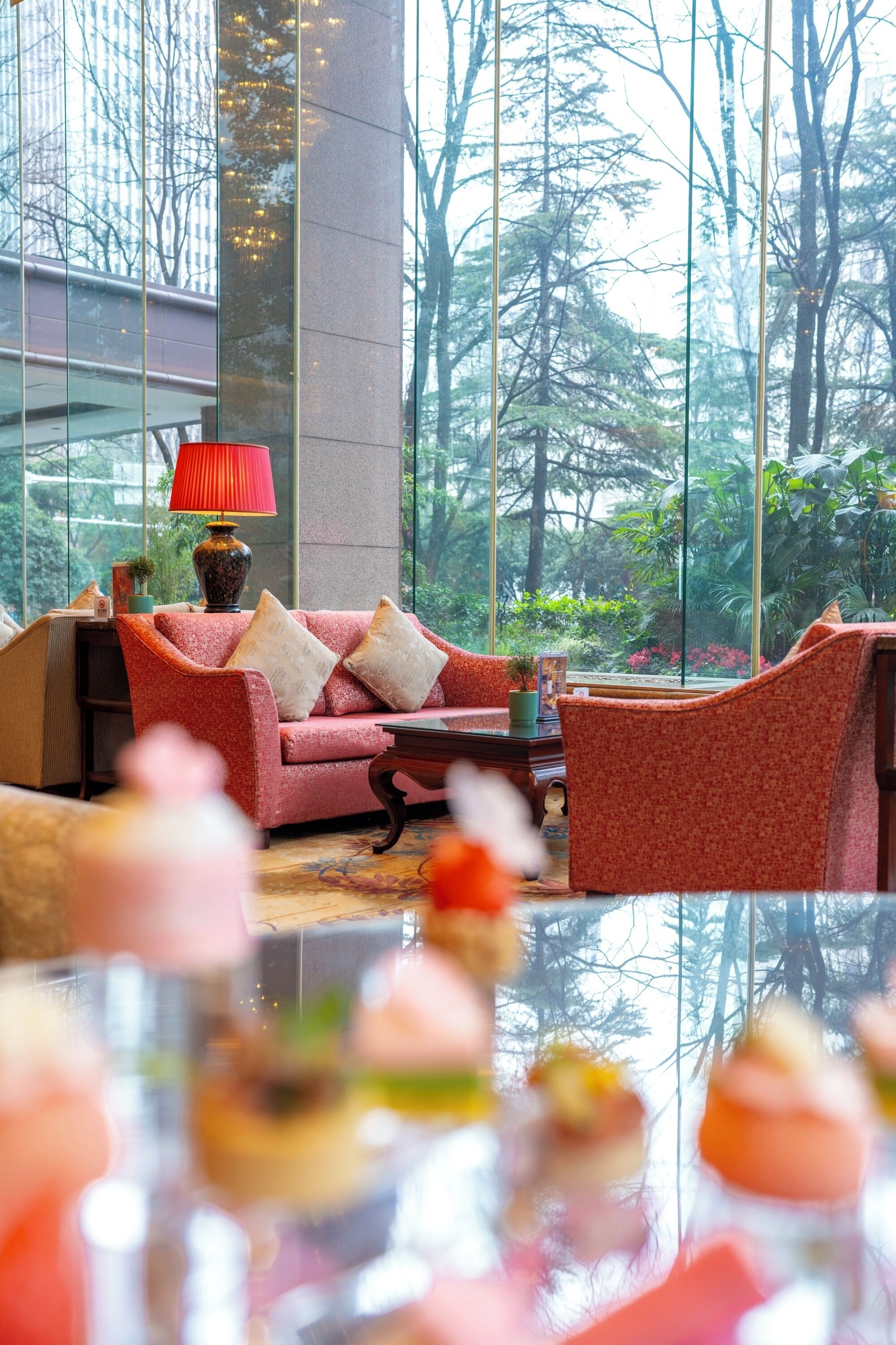 A modern hotel lobby with a red sofa, a coffee table, and a large window offering a view of trees and a garden.