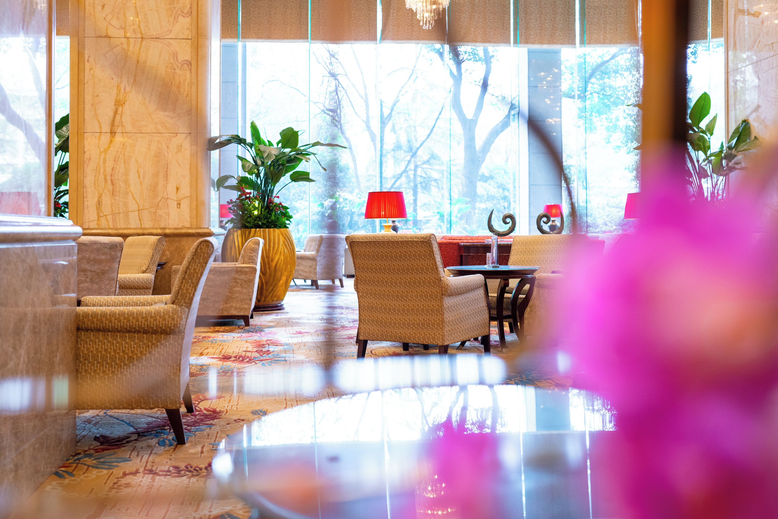 A hotel lobby with a marble reception desk, wicker chairs, a red lamp, and a large window with a view of trees.