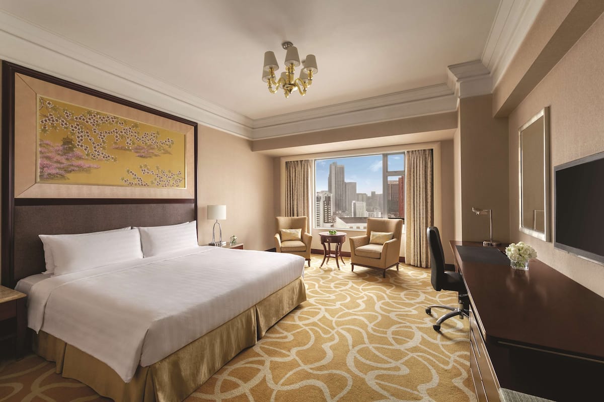 A hotel room with a large bed, a desk, two chairs, a chandelier, and a city view.