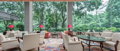 A spacious lobby with a large window offering a view of lush greenery, multiple seating areas with armchairs and sofas, and a patterned carpet.