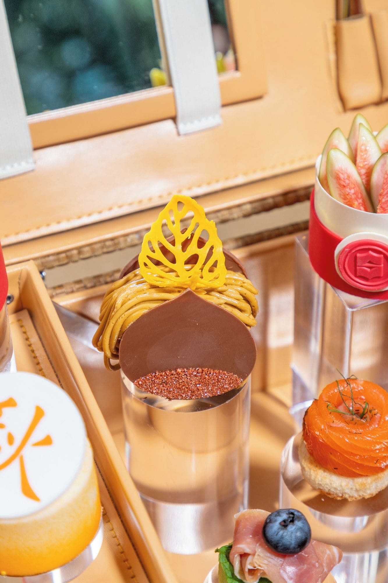 Una bandeja con diferentes alimentos, incluyendo un postre de chocolate con un adorno amarillo, un vaso con borde rojo y un plato de salmón y arándanos.