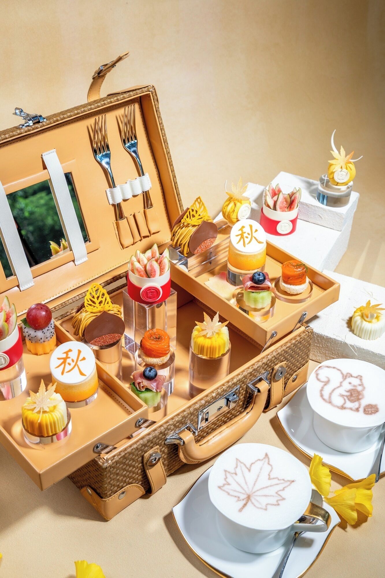 A collection of food items and utensils arranged on a table, including a suitcase and a tray with various desserts and a cup of coffee.