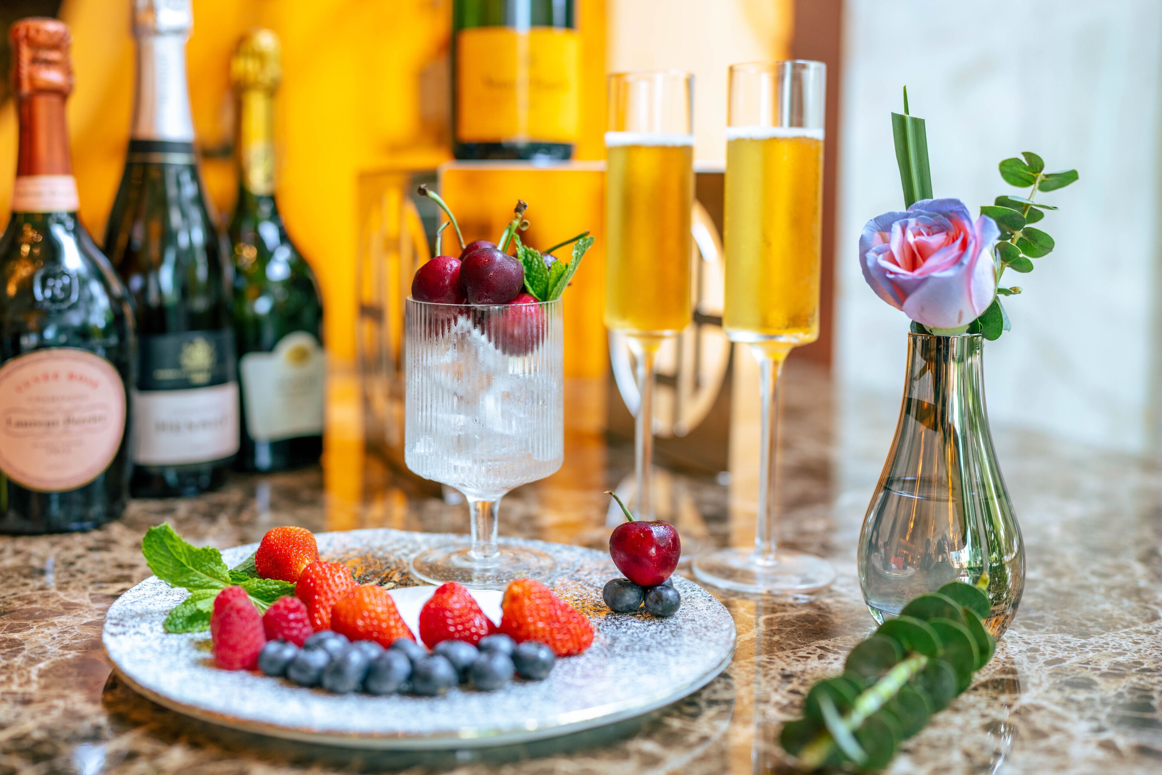 A table with a plate of strawberries and blueberries, a glass with a cherry garnish, champagne bottles, and glasses of beer.