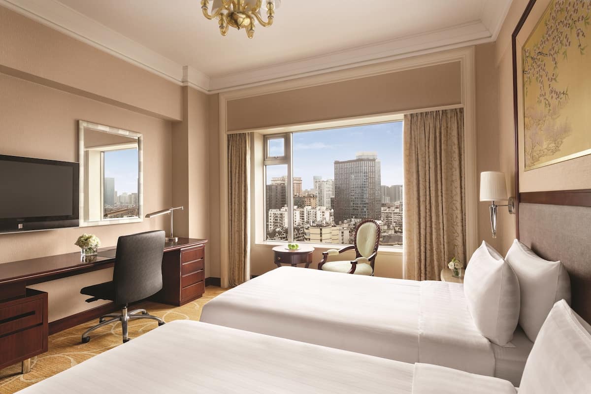 A hotel room with two beds, a desk, a chair, a television, and a large window with a city view.