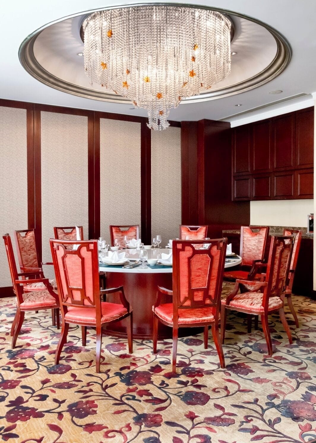 A dining room with a round table, red upholstered chairs, a chandelier, and a patterned carpet.