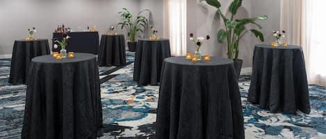 A conference room with round tables covered with black cloths, each with a candle and flowers. There are potted plants and a bar area with bottles.